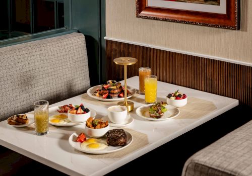 A dessert and breakfast spread on a booth table: assorted pastries, fruit bowls, pancakes, eggs, bacon, and several drinks including orange juice.