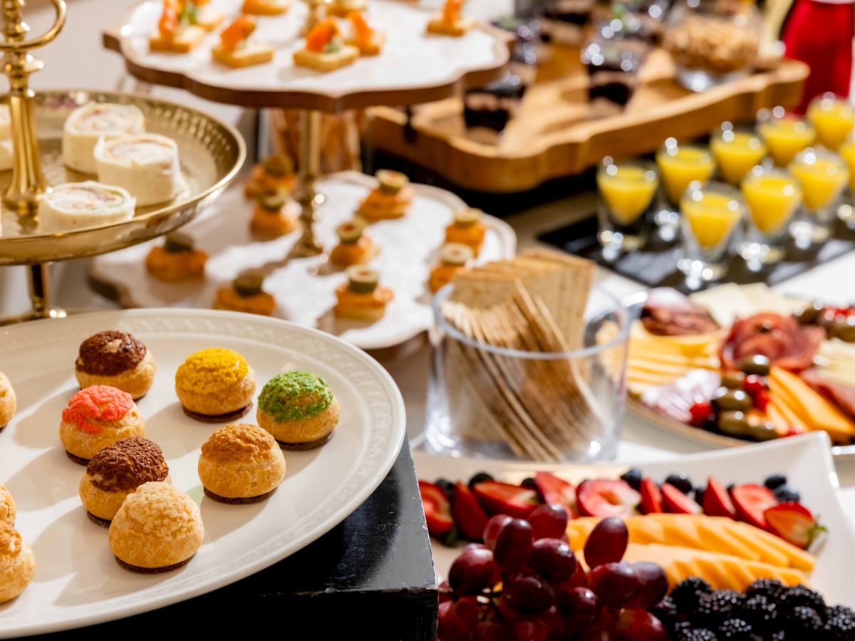 A catered buffet spread shows colorful mini pastries, fruit and veggie trays, and juice glasses on tiered serving stands.