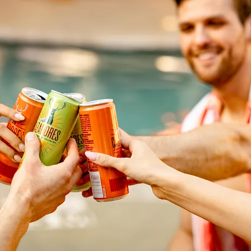 A group clinks colorful canned drinks outdoors by the water, smiling as they toast together.