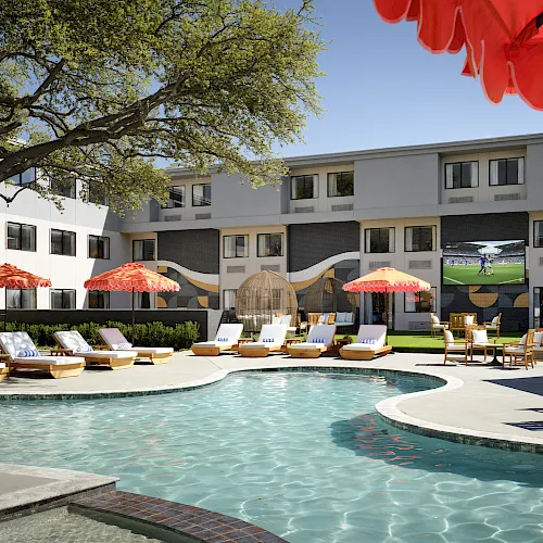 A hotel courtyard with a turquoise swimming pool, lounge chairs, red umbrellas, and a sunny multi-story building behind it.