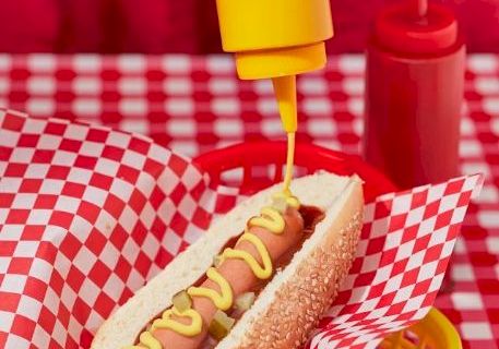 A hand squeezes yellow condiment onto a hot dog in checkered paper, with red trays and another drink in the background.