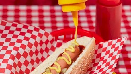 A hand squeezes yellow condiment onto a hot dog in checkered paper, with red trays and another drink in the background.